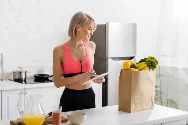 Mujer rubia alegre en ropa deportiva sosteniendo tableta digital y mirando bolsa de papel con comestibles - foto de stock