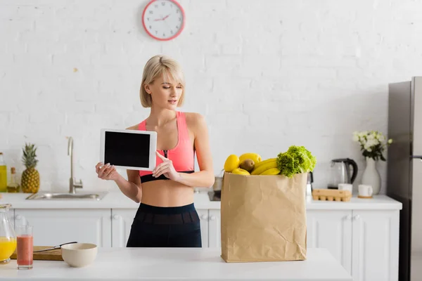 Schöne blonde Frau in Sportbekleidung hält digitale Tablette mit leerem Bildschirm in der Nähe Papiertüte mit Lebensmitteln — Stockfoto