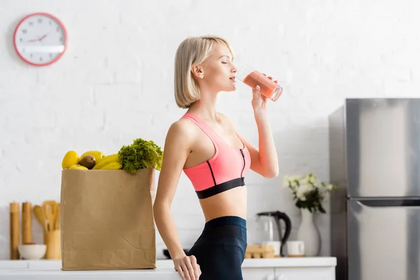 Schöne blonde Frau in Sportbekleidung trinkt Smoothie in der Nähe einer Papiertüte mit Lebensmitteln — Stockfoto