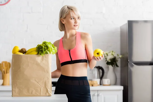Attractive blonde woman in sportswear holding apple near paper bag with groceries — Stock Photo