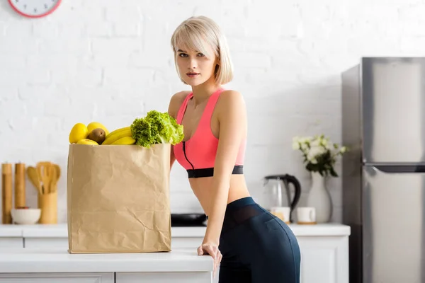Blonde woman in sportswear looking at camera near paper bag with groceries — Stock Photo
