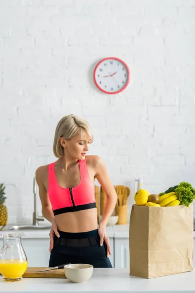 Mujer rubia en ropa deportiva mirando bolsa de papel con comestibles en la cocina - foto de stock