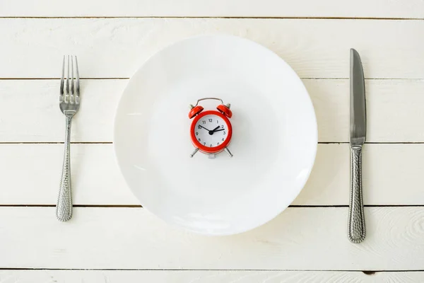 Top view of white plate with red alarm clock near cutlery on wooden surface — Stock Photo