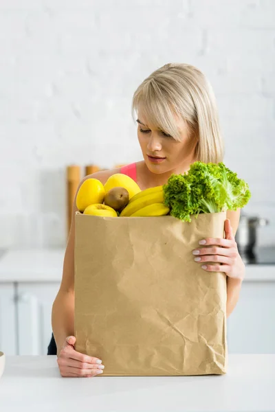 Femme blonde regardant sac en papier avec épicerie dans la cuisine — Photo de stock