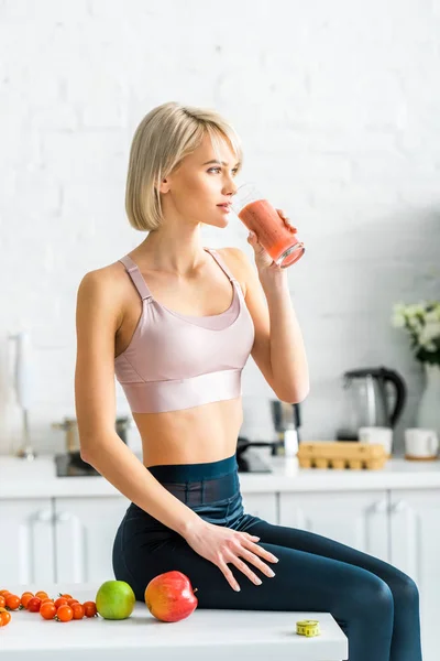 Atractiva chica rubia en ropa deportiva bebiendo sabroso smoothie mientras está sentado en la cocina cerca de manzanas - foto de stock