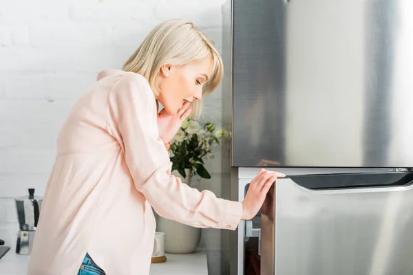 Surprised blonde pregnant woman looking at fridge in kitchen — Stock Photo