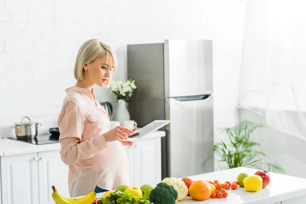 Blonde Schwangere nutzt digitales Tablet in der Nähe von Lebensmitteln in der Küche — Stockfoto