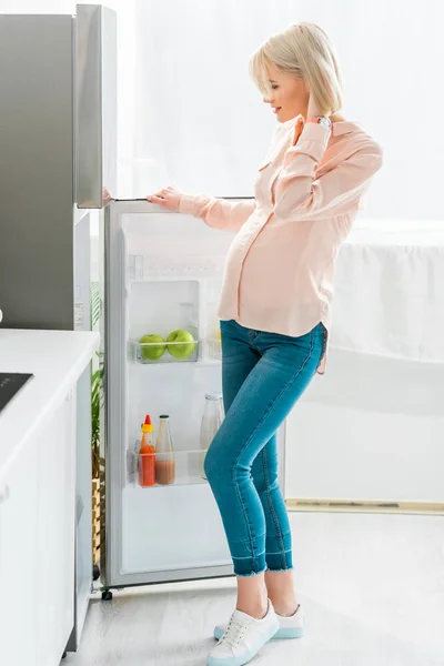 Feliz mulher grávida loira em pé perto de geladeira na cozinha — Fotografia de Stock