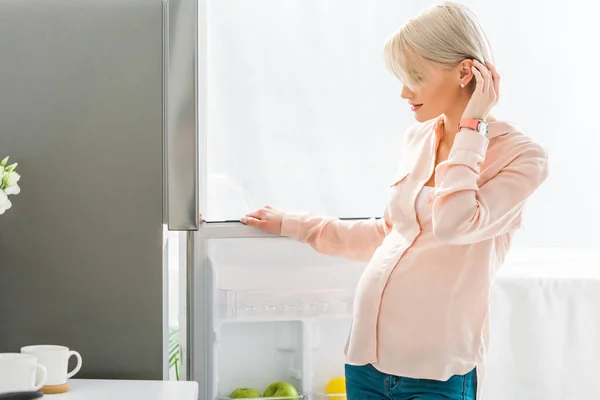 Glückliche blonde schwangere Frau berührt Haare, während sie in der Küche neben Kühlschrank steht — Stockfoto