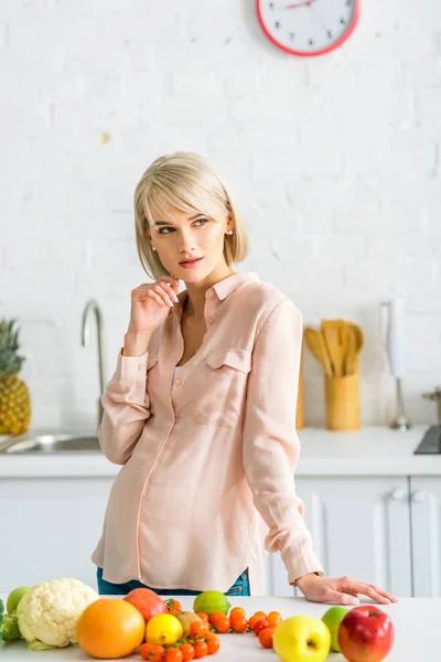 Pensive blonde pregnant woman standing near fruits and vegetables — Stock Photo