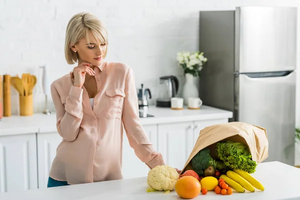 Blonde pregnant woman looking at tasty vegetables and fruits near paper bag — Stock Photo