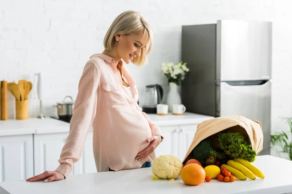 Atractiva mujer rubia y embarazada mirando comestibles cerca de bolsa de papel - foto de stock