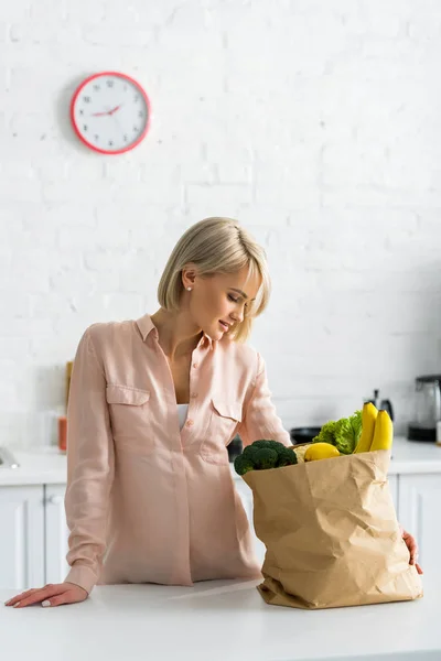 Jolie blonde, femme enceinte regardant sac en papier dans la cuisine — Photo de stock