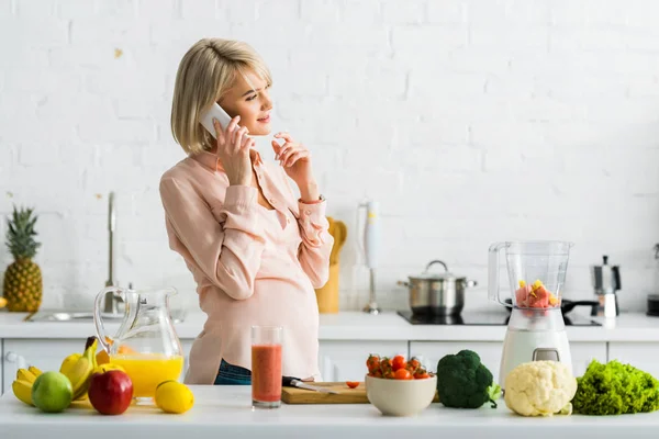 Blonde pregnant woman talking on smartphone in kitchen — Stock Photo