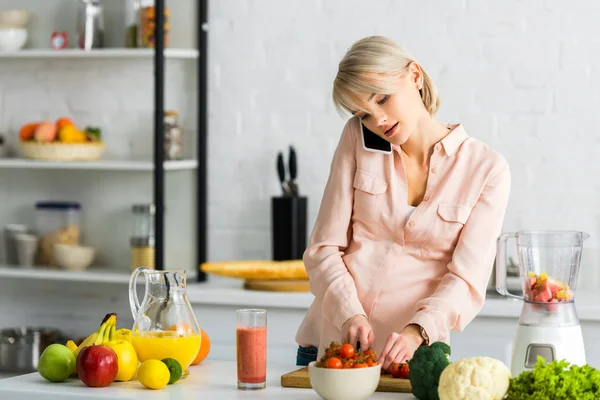 Mujer embarazada rubia hablando en el teléfono inteligente en la cocina cerca de frutas y verduras - foto de stock
