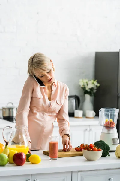 Mujer embarazada rubia hablando en el teléfono inteligente en la cocina cerca de tomates cherry en la tabla de cortar - foto de stock