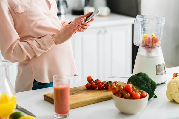 Обрезанный вид беременной женщины с помощью смартфона на кухне рядом с помидорами черри на разделочной доске — стоковое фото