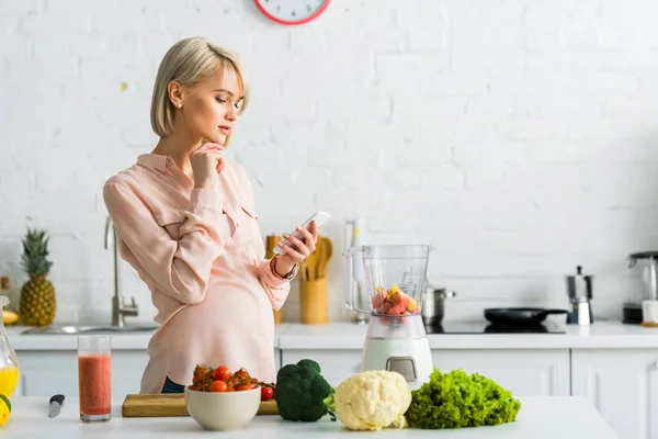 Mujer embarazada rubia usando teléfono inteligente en la cocina cerca de tomates cherry en la tabla de cortar - foto de stock