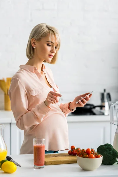 Donna incinta utilizzando smartphone in cucina vicino ai pomodorini ciliegini sul tagliere — Foto stock