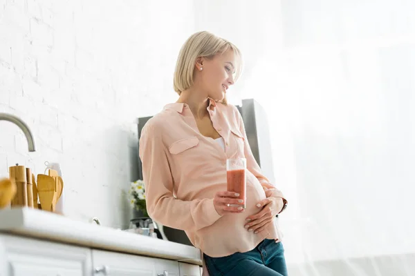 Cheerful blonde pregnant woman holding glass of smoothie in kitchen — Stock Photo