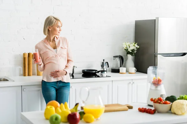 Blonde femme enceinte tenant verre de smoothie dans la cuisine — Photo de stock
