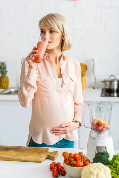 Atractiva mujer embarazada rubia bebiendo batido cerca de verduras en la cocina - foto de stock