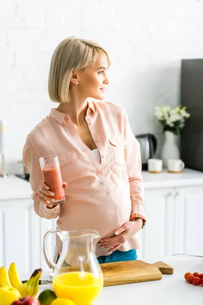 Feliz rubia embarazada joven de pie con un vaso de batido en la cocina - foto de stock