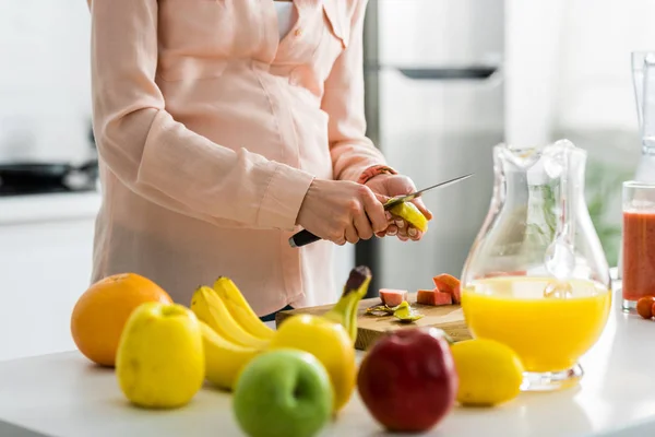 Vista cortada de grávida jovem mulher corte de frutas na cozinha — Fotografia de Stock