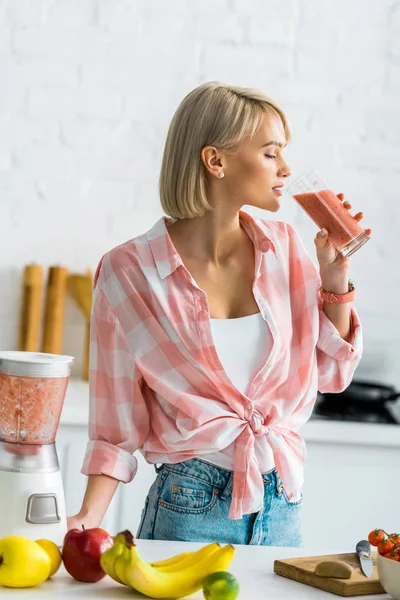 Attractive blonde woman drinking tasty smoothie in kitchen — Stock Photo