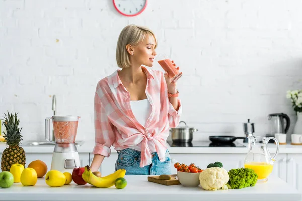 Mulher loira atraente beber smoothie saboroso perto de ingredientes na cozinha — Fotografia de Stock