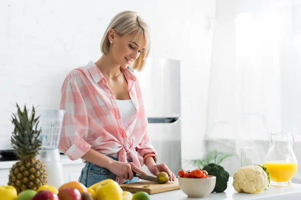 Mulher loira feliz corte kiwi frutas perto de ingredientes na cozinha — Fotografia de Stock