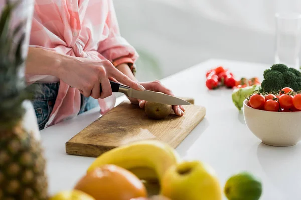 Cropped view of woman cutting kiwi fruit near ingredients in kitchen — Stock Photo