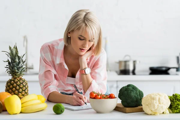 Mulher loira escrevendo em notebook perto de ingredientes na cozinha — Fotografia de Stock