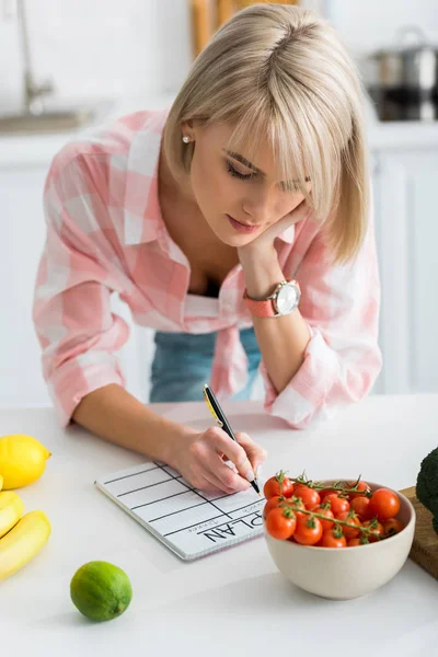 Atractiva mujer rubia escribiendo en cuaderno con letras de plan cerca de los ingredientes en la cocina - foto de stock