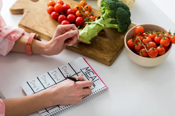 Vista cortada de menina escrevendo em caderno com refeição lettering perto de ingredientes na cozinha — Fotografia de Stock