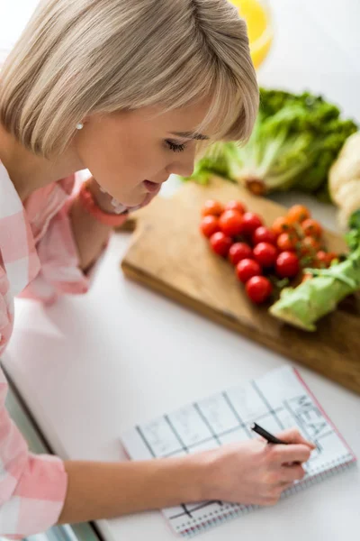 Vista aérea de niña escribiendo en cuaderno con letras de comida cerca de los ingredientes en la cocina - foto de stock