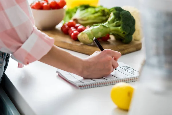 Vista ritagliata della donna che scrive nel taccuino con lettering pasto vicino ingredienti in cucina — Foto stock