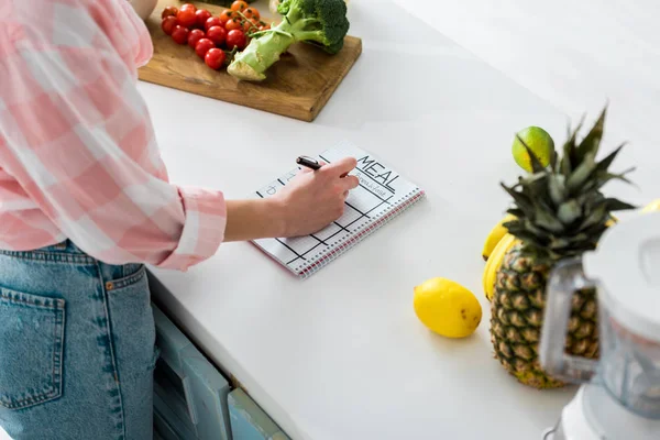 Vista cortada de menina escrevendo em caderno com refeição lettering perto de ingredientes saborosos na cozinha — Fotografia de Stock