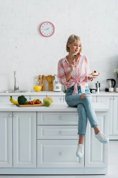 Atractiva chica rubia sentada cerca de frutas y verduras orgánicas y mirando la torta - foto de stock