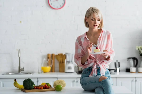 Блондинка сидить біля органічних фруктів і овочів і дивиться на торт — стокове фото
