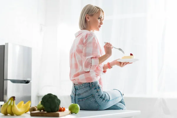 Блондинка держит вилку и блюдце со сладким тортом рядом с органическими фруктами и овощами — стоковое фото