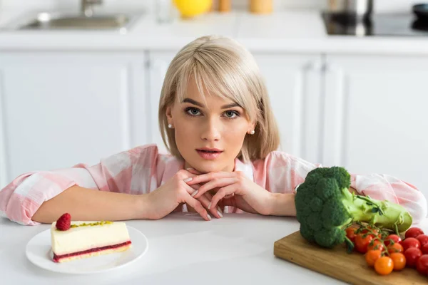 Blondes Mädchen blickt nahe Untertasse mit süßem Kuchen und Bio-Gemüse in die Kamera — Stockfoto