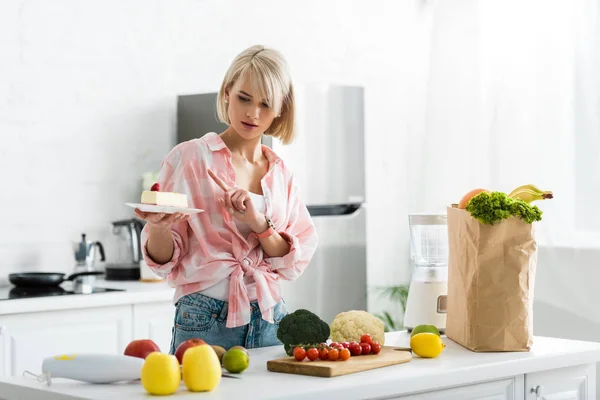 Chica rubia señalando con el dedo en platillo con pastel dulce cerca de verduras y frutas orgánicas - foto de stock