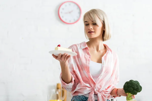 Blondes Mädchen blickt auf Untertasse mit süßem Kuchen, während sie Bio-Brokkoli in der Hand hält — Stockfoto