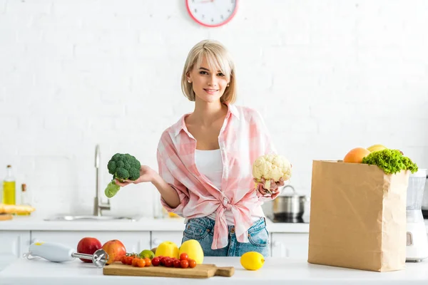 Cheerful blonde girl holding cauliflower and broccoli in kitchen — Stock Photo