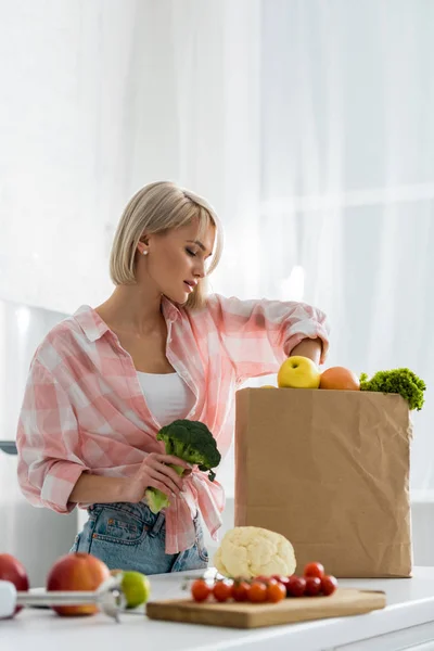 Attractive blonde girl holding organic broccoli near paper bag — Stock Photo
