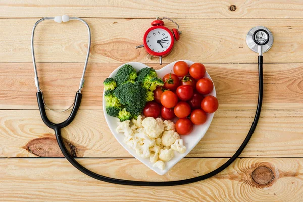 Top view of tasty vegetables on heart-shape plate near stethoscope and retro alarm clock on wooden surface — Stock Photo