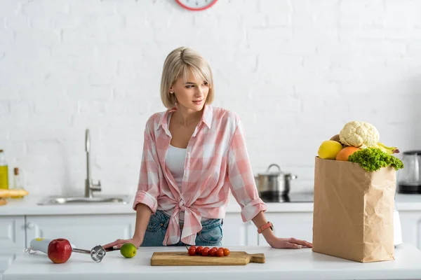 Mujer rubia de pie bolsa de papel con comestibles en la cocina - foto de stock