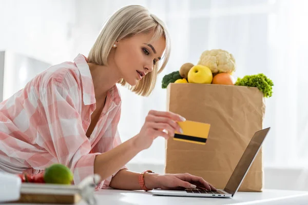 Atractiva chica rubia sosteniendo la tarjeta de crédito mientras usa el ordenador portátil cerca de la bolsa de papel con comestibles - foto de stock