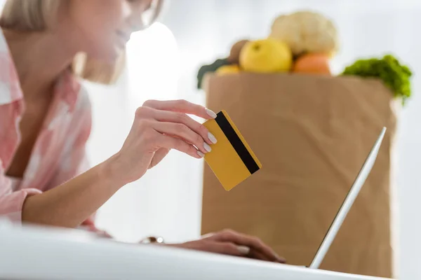 Обрізаний вигляд дівчина тримає кредитну картку при використанні ноутбука біля паперового пакета з продуктами харчування — стокове фото
